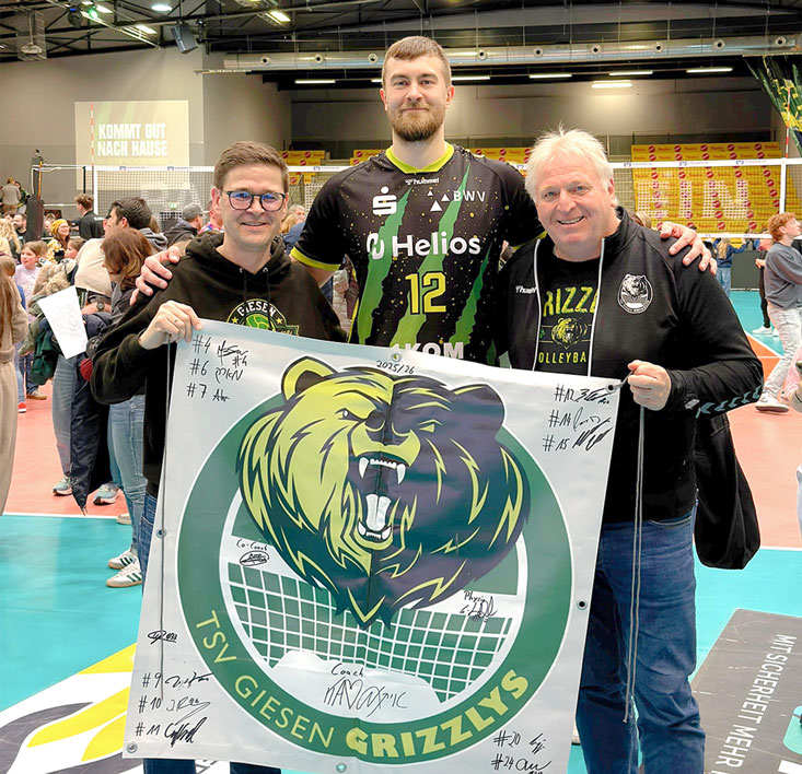 Ralf Köster (rechts) hat mit seinem Banner fast alle Hallen und Arenen der Volleyball-Bundesligisten bereist. Auch Mittelblocker Jakob Günthör hat sich wie seine Teamkollegen mit seiner Unterschrift darauf verewigt. Unterstützung bei der Aktion gibt es nun von Oliver Kroll und dem Gleitz Verlag. Alle Beteiligten wünschen sich einen möglichst hohen Erlös für die Clinic-Clowns Hannover. Foto: Lukas Piel